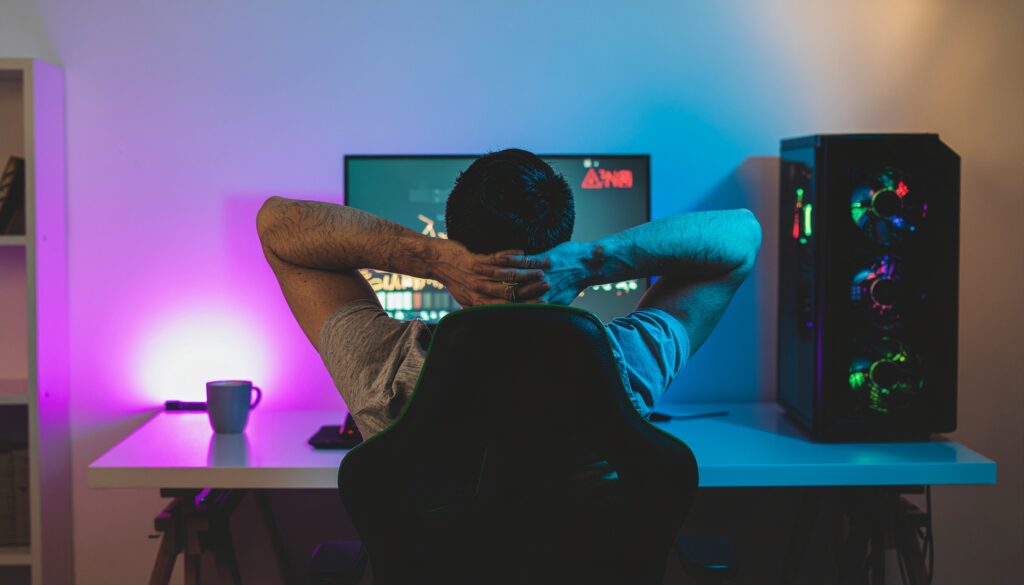 青や紫のLEDに照らされた部屋で、警告アイコンが表示されたPCモニターの前で腕を組み、落ち着いて待機しているゲーマーのイラスト。